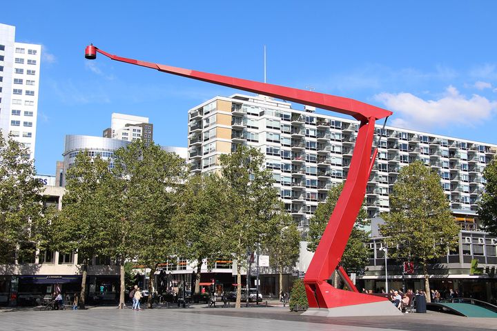One of the tall red lamps on Schouwburgplein, with trees and blocks of flats behind it.